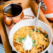 Bowl of chili with cheese, corn chips, and sour cream. Copper cup of cider. All on wooden tray.